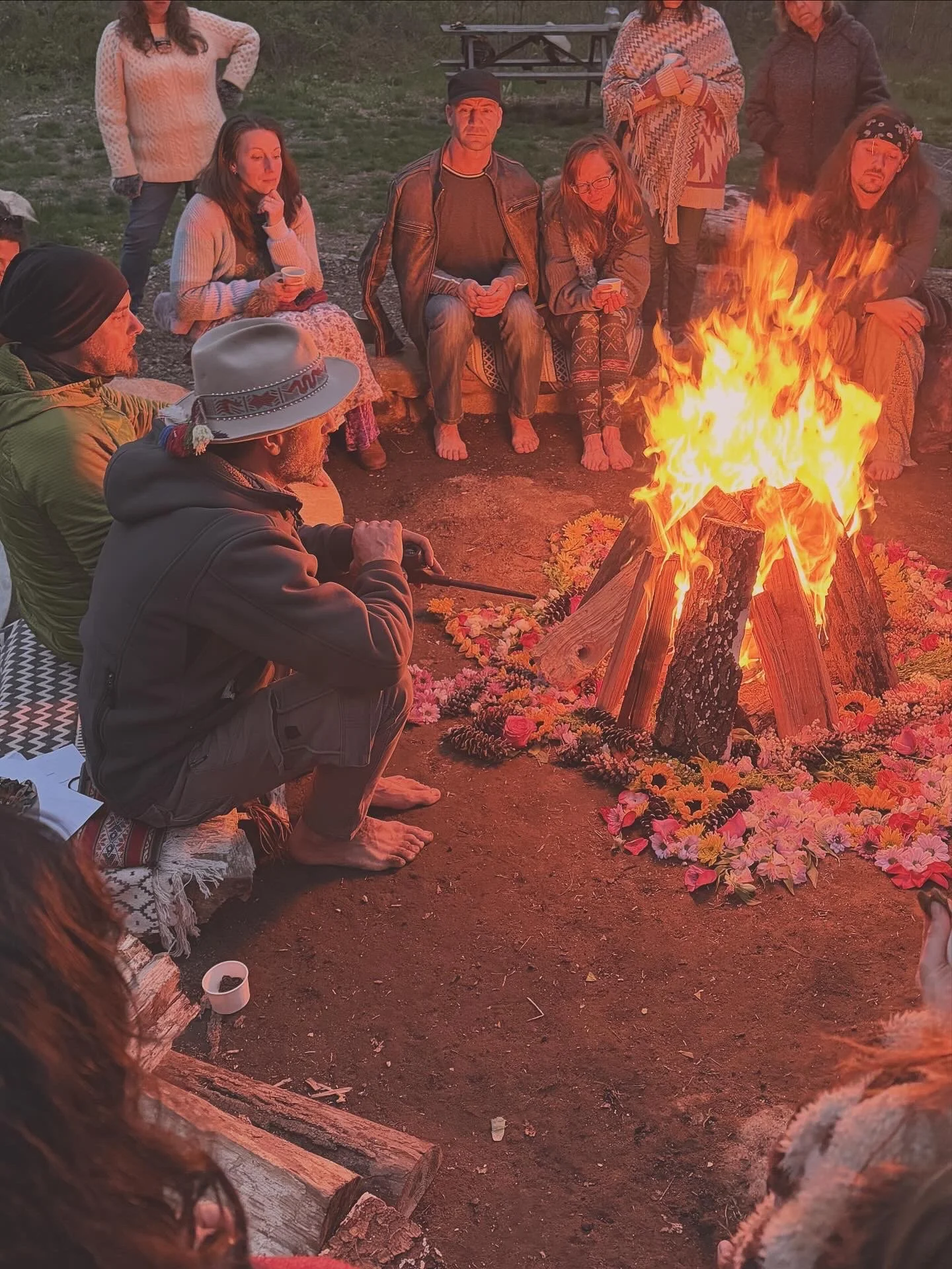 People sit around a dusk bonfire ringed by a colorful flower-and-pinecone mandala