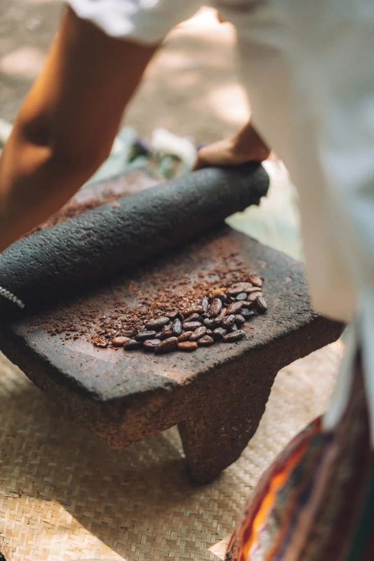 Hands grind whole roasted cacao beans on a dark stone metate