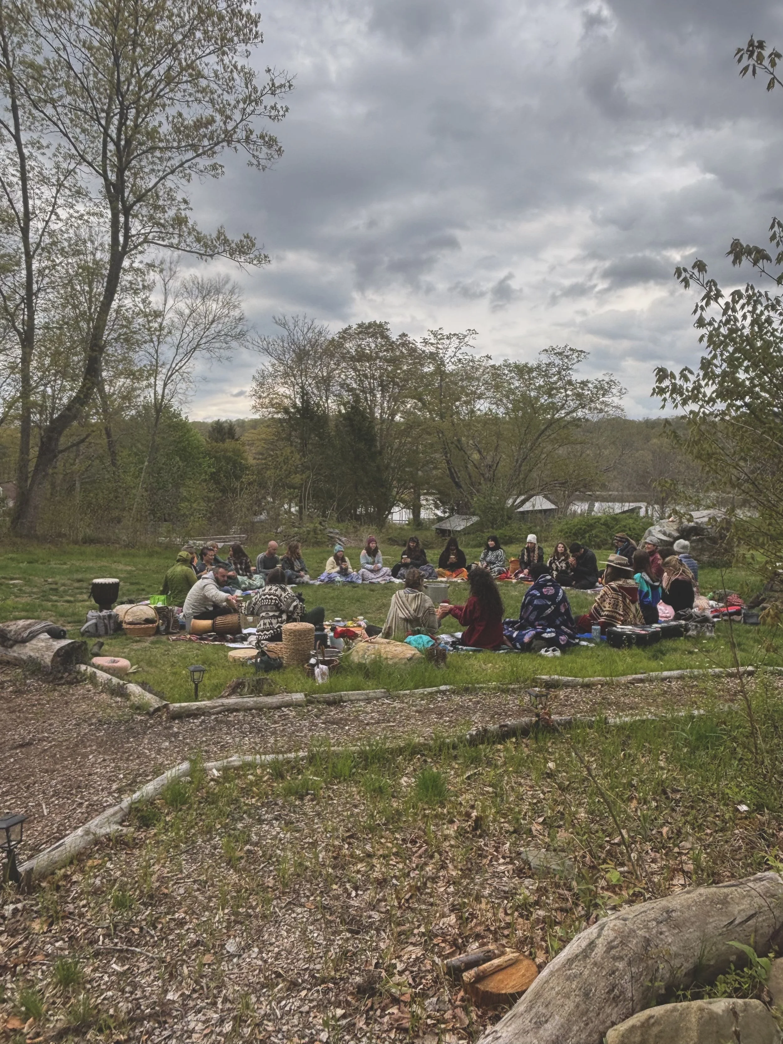 A large group sits in a circle on blankets in a grassy field for an outdoor cacao ceremony