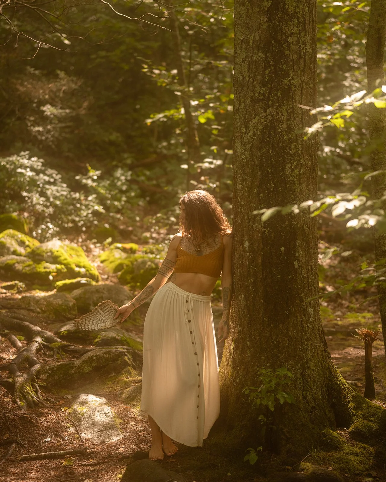 Victoria Moon stands barefoot against a forest tree holding a patterned feather