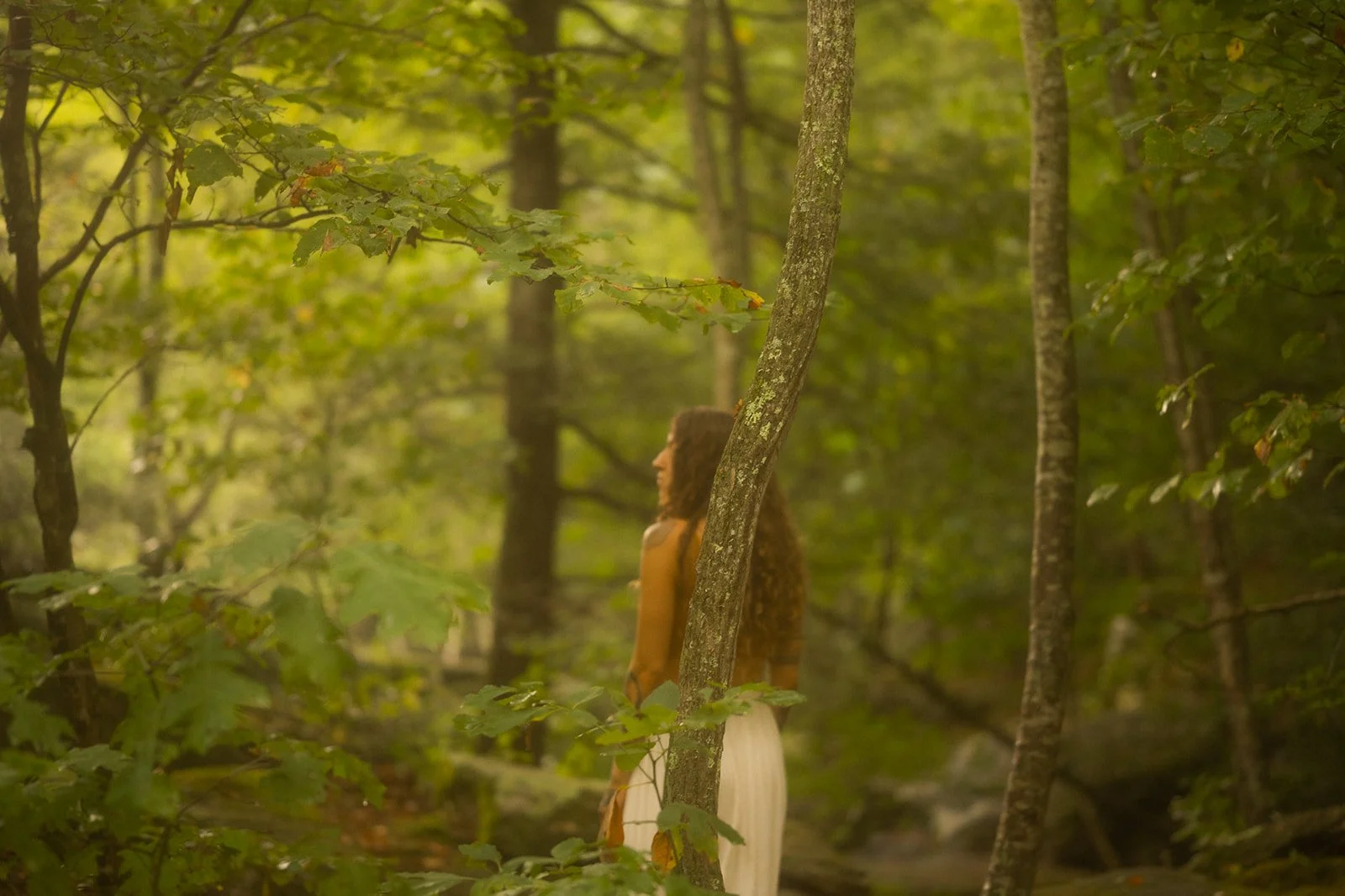 Victoria Moon stands in profile in a sunlit forest in a mustard sleeveless top and long white skirt